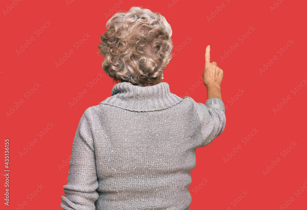 Senior grey-haired woman wearing casual winter sweater posing backwards pointing ahead with finger hand