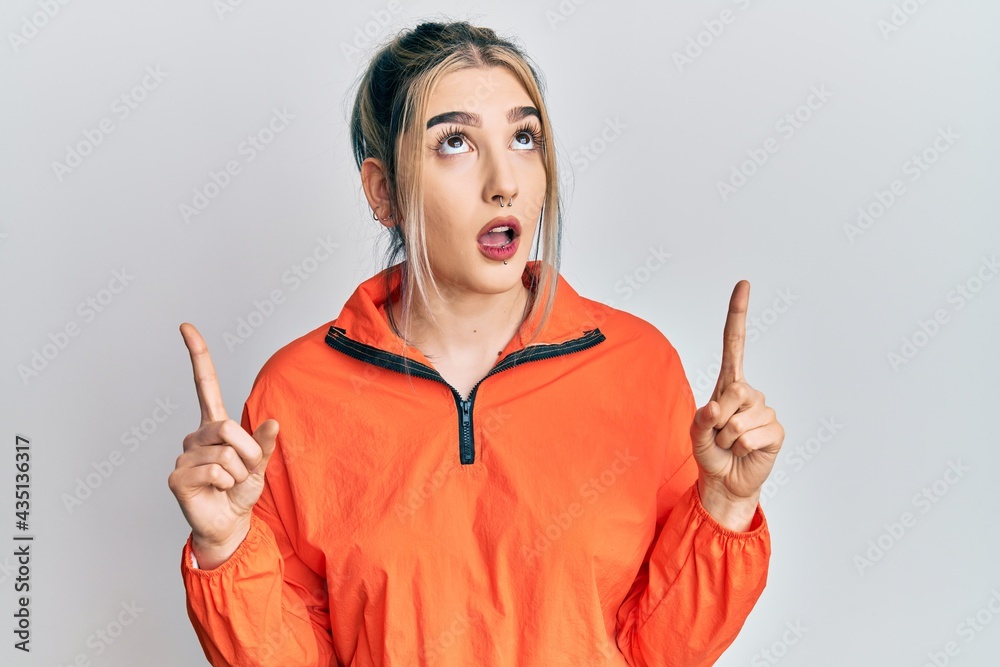 Young modern girl wearing sports sweatshirt amazed and surprised looking up and pointing with fingers and raised arms. Young modern girl wearing sports sweatshirt amazed and surprised looking up and pointing with fingers and raised arms.