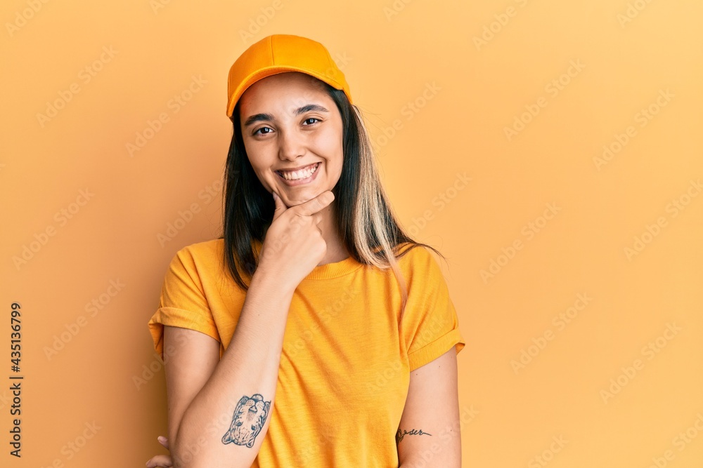 Young hispanic woman wearing delivery uniform and cap looking confident at the camera smiling with crossed arms and hand raised on chin. thinking positive.