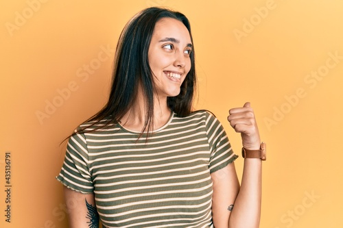 Young hispanic woman wearing casual striped t shirt smiling with happy face looking and pointing to the side with thumb up.