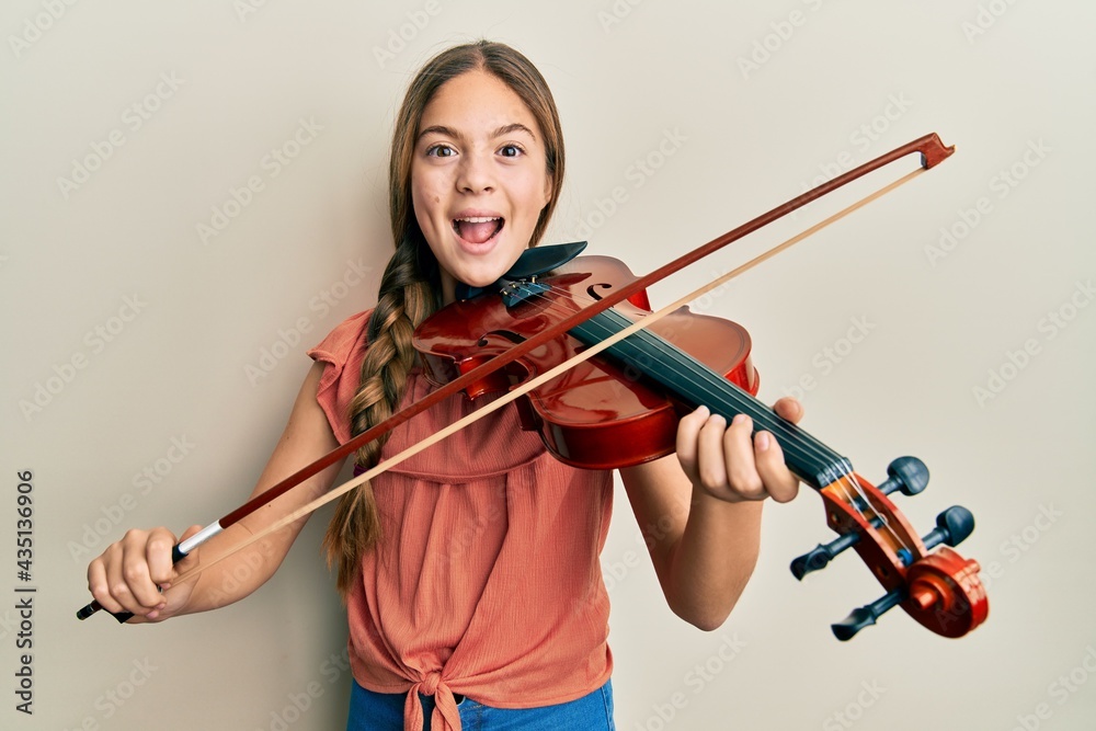 Beautiful brunette little girl playing violin celebrating crazy and amazed for success with open eyes screaming excited. Beautiful brunette little girl playing violin celebrating crazy and amazed for success with open eyes screaming excited.