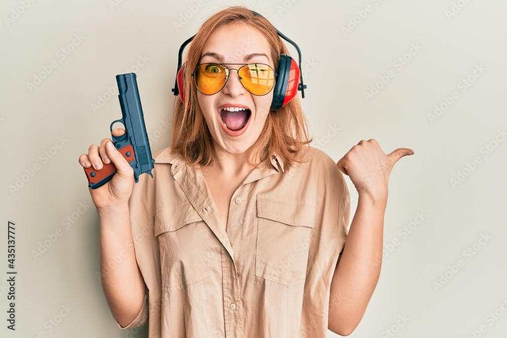 Young caucasian shooter woman using gun pointing thumb up to the side smiling happy with open mouth