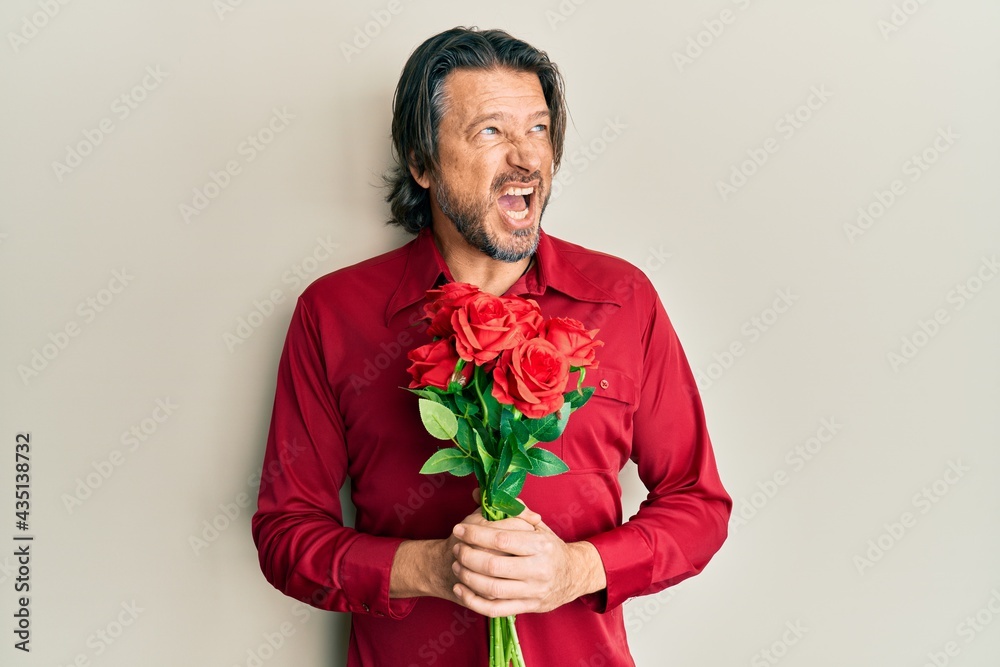 Middle age handsome man holding bouquet of red roses angry and mad screaming frustrated and furious, shouting with anger. rage and aggressive concept. Middle age handsome man holding bouquet of red roses angry and mad screaming frustrated and furious, shouting with anger. rage and aggressive concept.