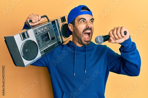 Young hispanic man holding boombox, listening to music singing with microphone angry and mad screaming frustrated and furious, shouting with anger. rage and aggressive concept.