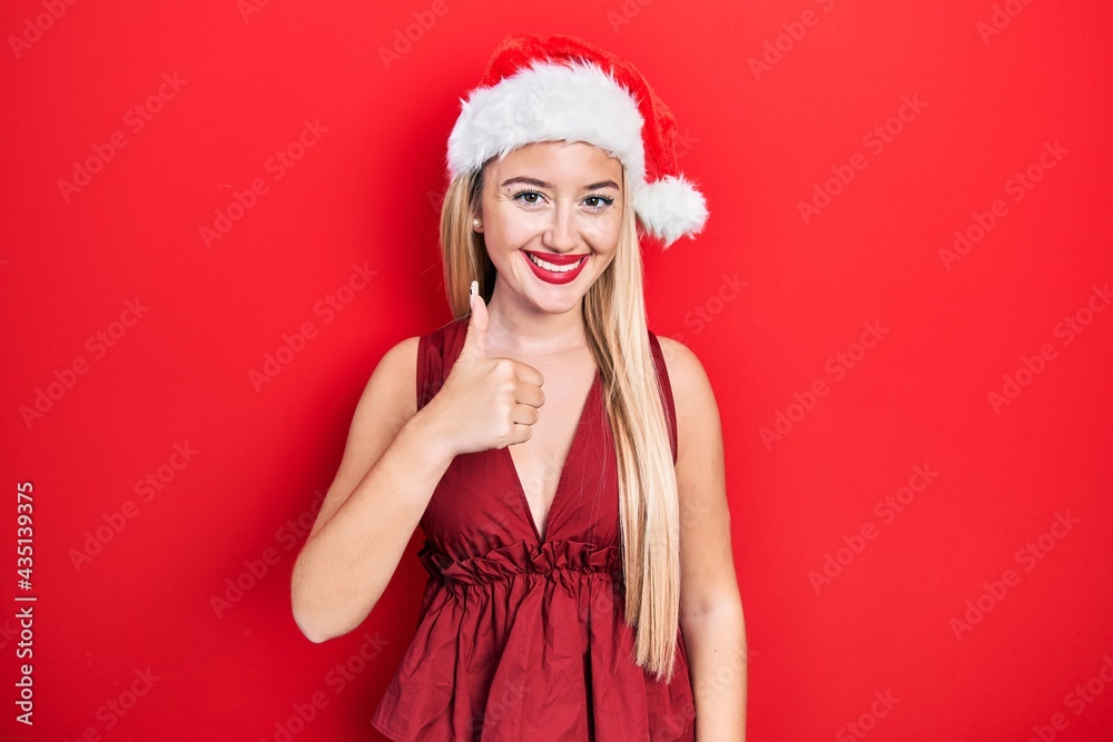 Young blonde girl wearing christmas hat doing happy thumbs up gesture with hand. approving expression looking at the camera showing success. Young blonde girl wearing christmas hat doing happy thumbs up gesture with hand. approving expression looking at the camera showing success.
