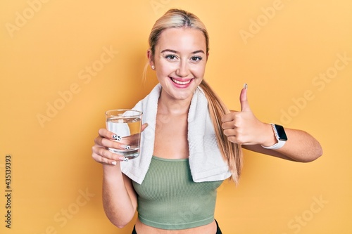Young blonde girl wearing sportswear drinking glass of water smiling happy and positive, thumb up doing excellent and approval sign © Krakenimages.com