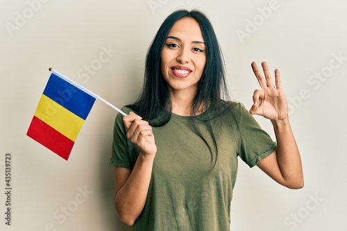Young hispanic girl holding romania flag doing ok sign with fingers, smiling friendly gesturing excellent symbol