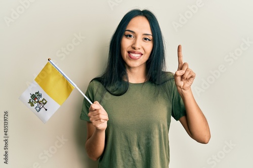 Young hispanic girl holding vatican city flag smiling with an idea or question pointing finger with happy face, number one