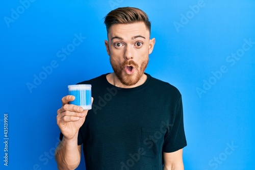 Young redhead man holding earwax cotton remover scared and amazed with open mouth for surprise, disbelief face