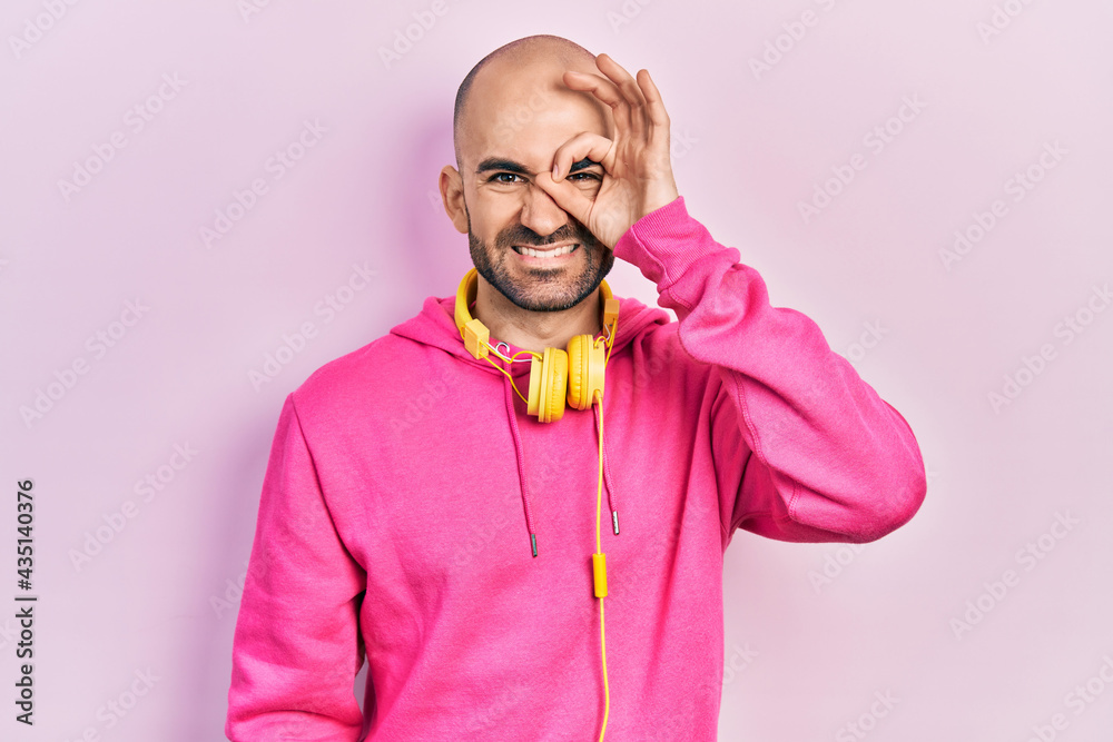 Fototapeta premium Young bald man wearing gym clothes and using headphones doing ok gesture with hand smiling, eye looking through fingers with happy face.