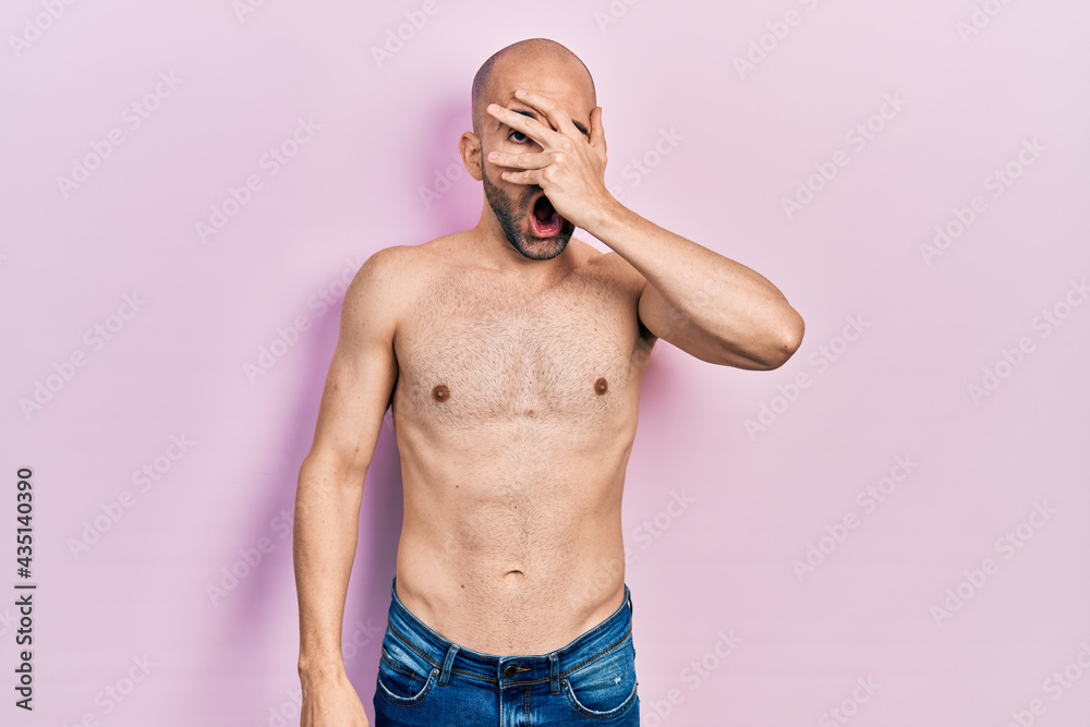 Fototapeta premium Young bald man standing shirtless peeking in shock covering face and eyes with hand, looking through fingers with embarrassed expression.