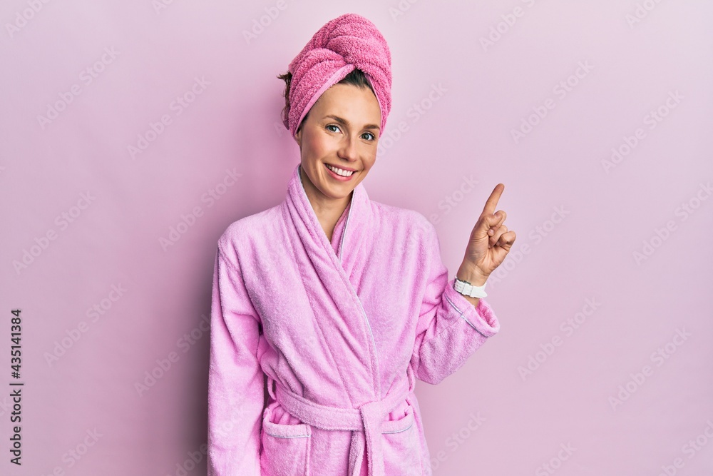Young blonde woman wearing shower towel cap and bathrobe with a big smile on face, pointing with hand finger to the side looking at the camera. Young blonde woman wearing shower towel cap and bathrobe with a big smile on face, pointing with hand finger to the side looking at the camera.