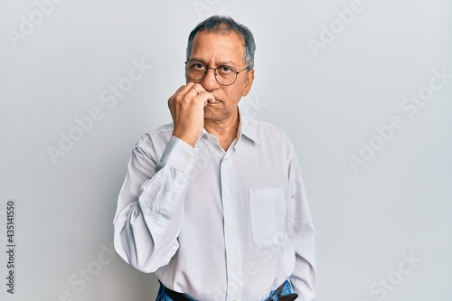 Middle age indian man wearing casual clothes and glasses looking stressed and nervous with hands on mouth biting nails. anxiety problem.