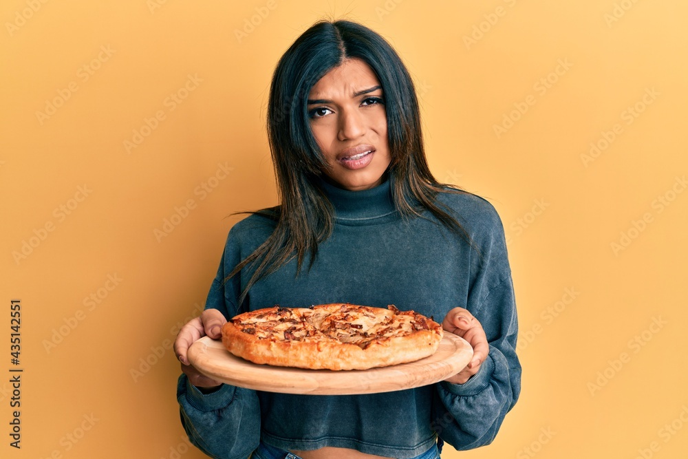 Young latin transsexual transgender woman holding italian pizza clueless and confused expression. doubt concept.
