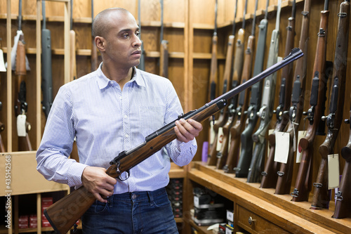 Portrait of interested hispanic man carefully choosing hunting rifle in modern gun shop