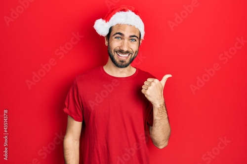 Young hispanic man wearing christmas hat smiling with happy face looking and pointing to the side with thumb up.