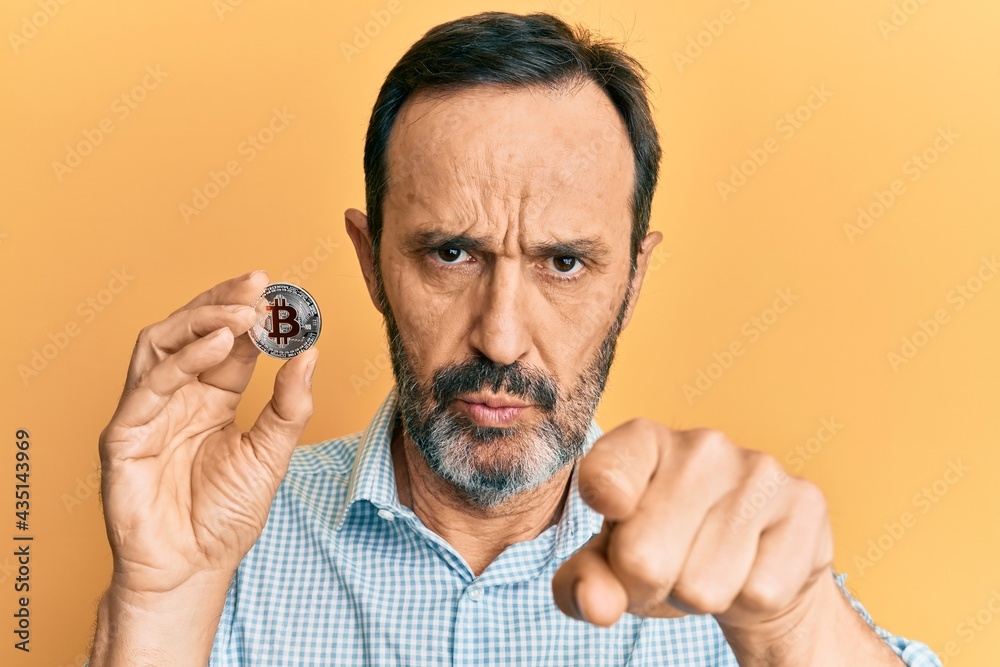 Middle age hispanic man holding virtual currency bitcoin pointing with finger to the camera and to you, confident gesture looking serious Middle age hispanic man holding virtual currency bitcoin pointing with finger to the camera and to you, confident gesture looking serious