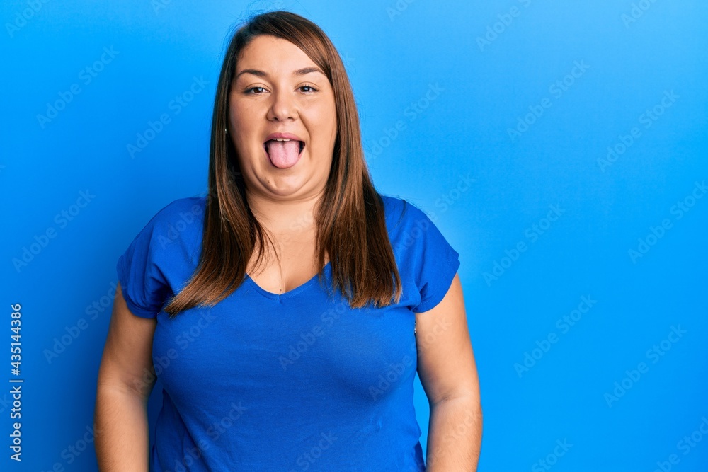 Beautiful brunette plus size woman wearing casual blue t shirt sticking tongue out happy with funny expression. emotion concept.