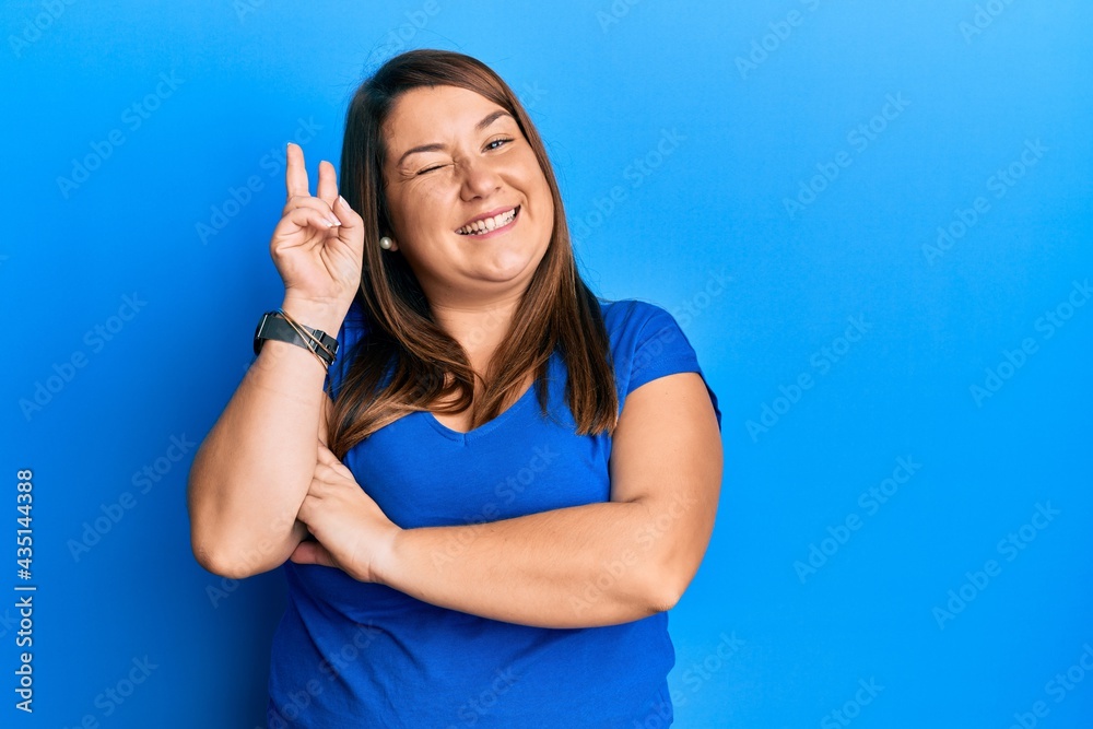 Fototapeta premium Beautiful brunette plus size woman wearing casual blue t shirt smiling with happy face winking at the camera doing victory sign. number two.