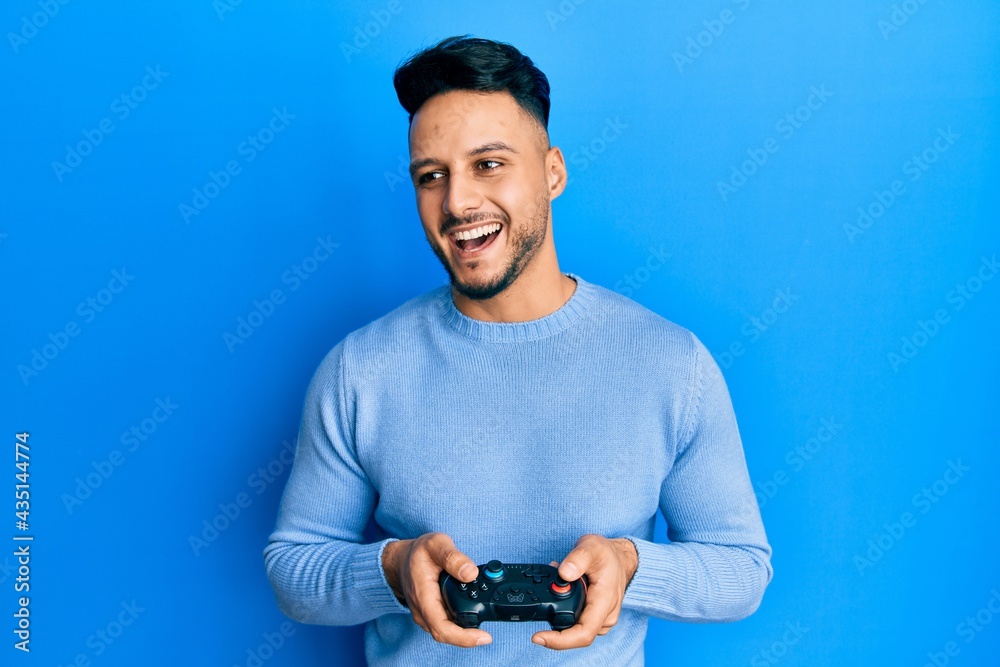 Young arab man playing video game holding controller smiling and laughing hard out loud because funny crazy joke.