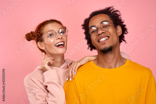Cuadro en lienzo Portrait of cheerful mixed race couple in love with glasses of positive dark skinned guy has date with red haired girlfriend, have fun together, pose at camera with broad smiles on pink