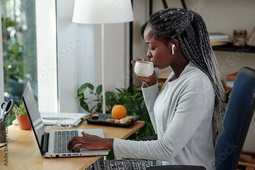 Young businesswoman drinking cup of tea or coffee and reading e-mails on laptop screen photo