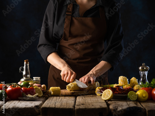 In the photo, a chef is cutting vegetables on a cutting board. There are many bright colorful ingredients on the wooden table - vegetables, spices, sliced lemon. Dark tones.
