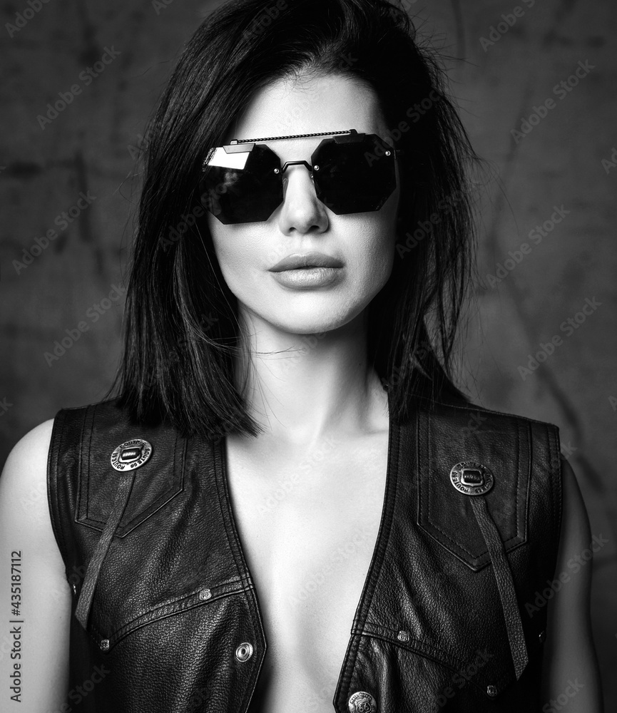 Portrait of young sexy pretty lady in black leather jacket and sunglasses standing and looking at camera over grey background. Beautiful women and sexual outfit concept Portrait of young sexy pretty lady in black leather jacket and sunglasses standing and looking at camera over grey background. Beautiful women and sexual outfit concept