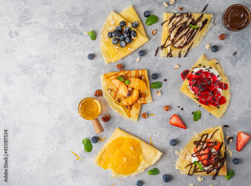 Variation of crepes or thin pancakes with fresh fruit, berries, cream cheese, honey, chocolate sauce on a gray concrete background. Top view, copy space.