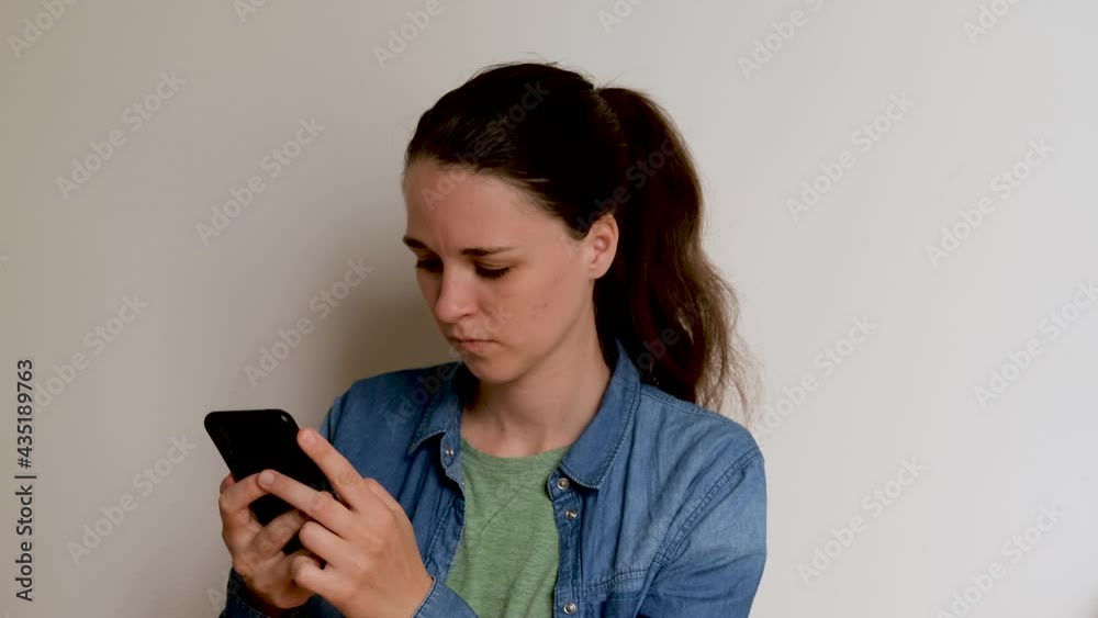 Young Caucasian European woman in shirt on white background is texting using smartphone and looking closely at phone screen. Important conversation and correspondence over Internet online. 4K footage. Young Caucasian European woman in shirt on white background is texting using smartphone and looking closely at phone screen. Important conversation and correspondence over Internet online. 4K footage.