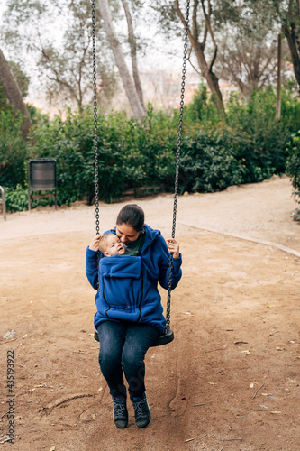 Mom in a blue warm sweater and jeans is swinging on a swing with a tiny baby on her chest
