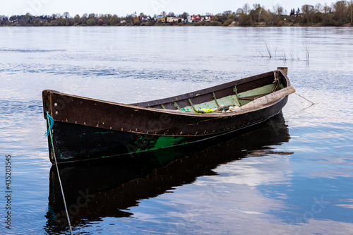 Small weathered wooden fishing row boat at coast on river  housing visible other side of coast. Brown green fishing vessel reflecting in blue calm water. Wooden boat ready to go.