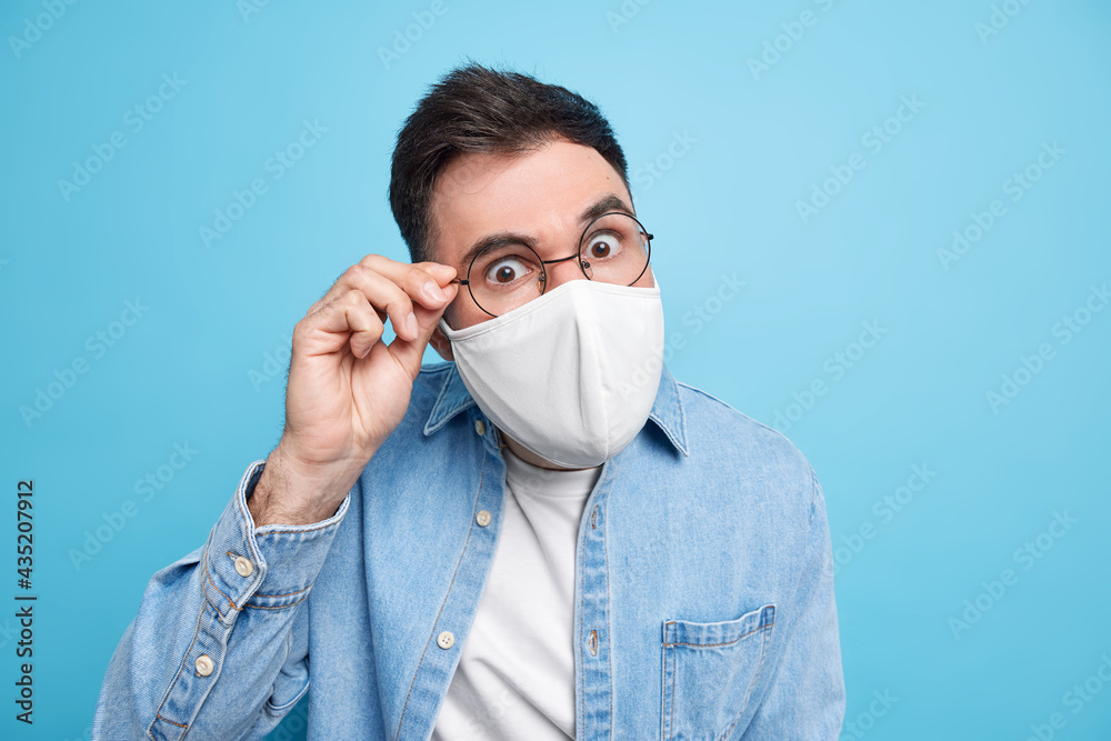 Covid 19 concept. Serious attentive adult man looks through spectacles wears protective face mask asks to follow social distancing rules dressed in denim shirt isolated over blue background. Covid 19 concept. Serious attentive adult man looks through spectacles wears protective face mask asks to follow social distancing rules dressed in denim shirt isolated over blue background.