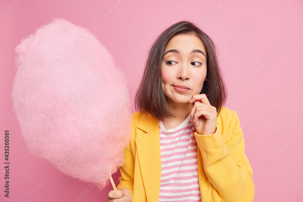 Studio shot of thoughtful young Asian woman looks at delicious candy floss hesitates whether to eat sweet dessert dressed in fashionable clothes isolated over pink background. What tasty dessert Studio shot of thoughtful young Asian woman looks at delicious candy floss hesitates whether to eat sweet dessert dressed in fashionable clothes isolated over pink background. What tasty dessert
