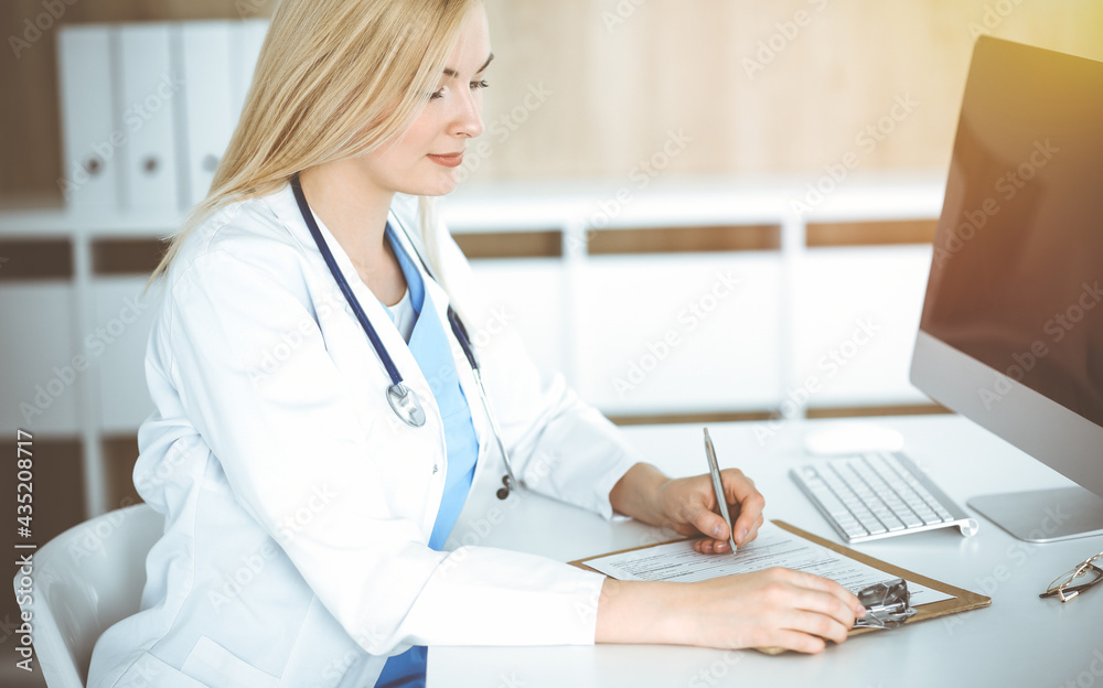 Woman-doctor at work while sitting at the desk in sunny clinic. Blonde cheerful physician filling up medication history record form