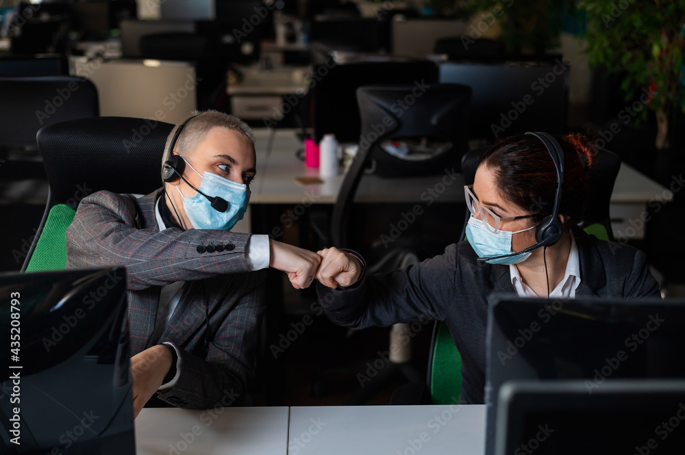 Two business women in masks are giving a high five while sitting at one desk in the office