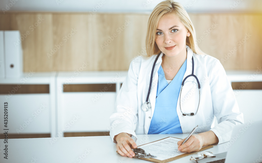 Woman-doctor at work while sitting at the desk in sunny clinic. Blonde cheerful physician filling up medication history record form Woman-doctor at work while sitting at the desk in sunny clinic. Blonde cheerful physician filling up medication history record form