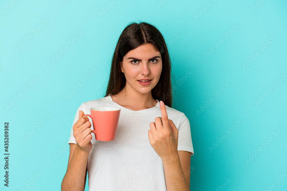 Young caucasian woman holding a mug isolated on blue background pointing with finger at you as if inviting come closer. Young caucasian woman holding a mug isolated on blue background pointing with finger at you as if inviting come closer.