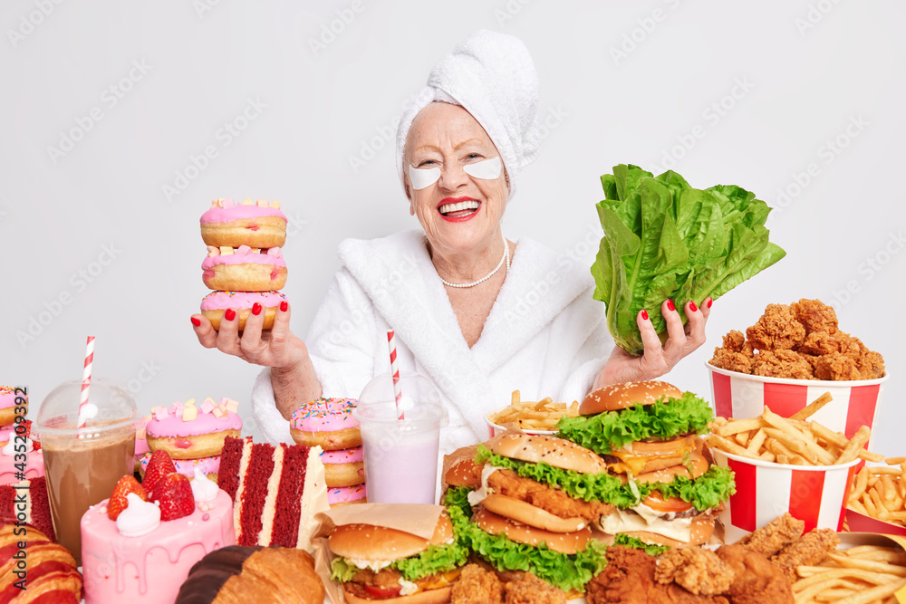 Positive old wrinkled woman smiles happily chooses between green lettuce salad and doughnuts has good mood surrounded by high calorie snacksapplies patches under eyes dressed in soft white bathrobe Positive old wrinkled woman smiles happily chooses between green lettuce salad and doughnuts has good mood surrounded by high calorie snacksapplies patches under eyes dressed in soft white bathrobe
