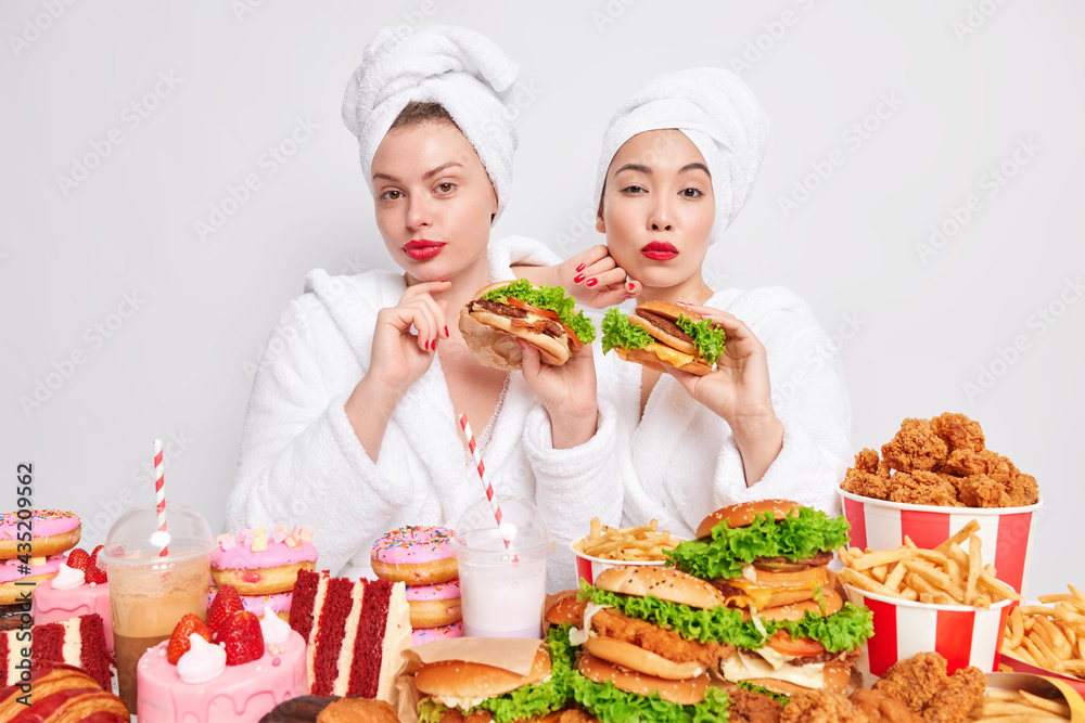 Binge eating habit concept. Two serious women wear red lipstick hold tasty hamburgers wear white soft bath dressing gowns and wrapped towels on head isolated over white background. Fast food eaters Binge eating habit concept. Two serious women wear red lipstick hold tasty hamburgers wear white soft bath dressing gowns and wrapped towels on head isolated over white background. Fast food eaters