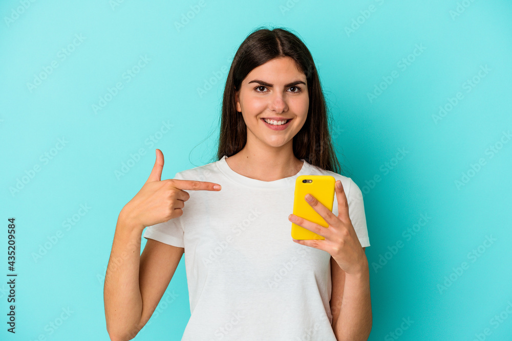Young caucasian woman holding a mobile phone isolated on blue background person pointing by hand to a shirt copy space, proud and confident Young caucasian woman holding a mobile phone isolated on blue background person pointing by hand to a shirt copy space, proud and confident