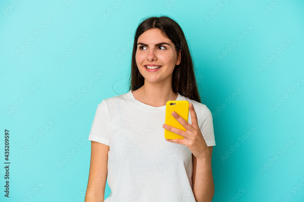 Young caucasian woman holding a mobile phone isolated on blue background dreaming of achieving goals and purposes Young caucasian woman holding a mobile phone isolated on blue background dreaming of achieving goals and purposes