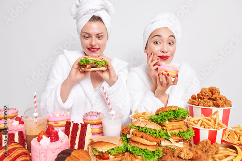 Horizontal shot of happy women enjoy domestic party hold hamburger and doughnut efford to eat junk food surrounded by many delicious snacks starve for cheat meal wear comfortable white bathrobes photo
