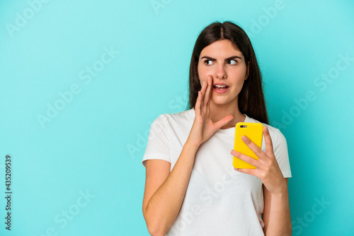 Young caucasian woman holding a mobile phone isolated on blue background is saying a secret hot braking news and looking aside