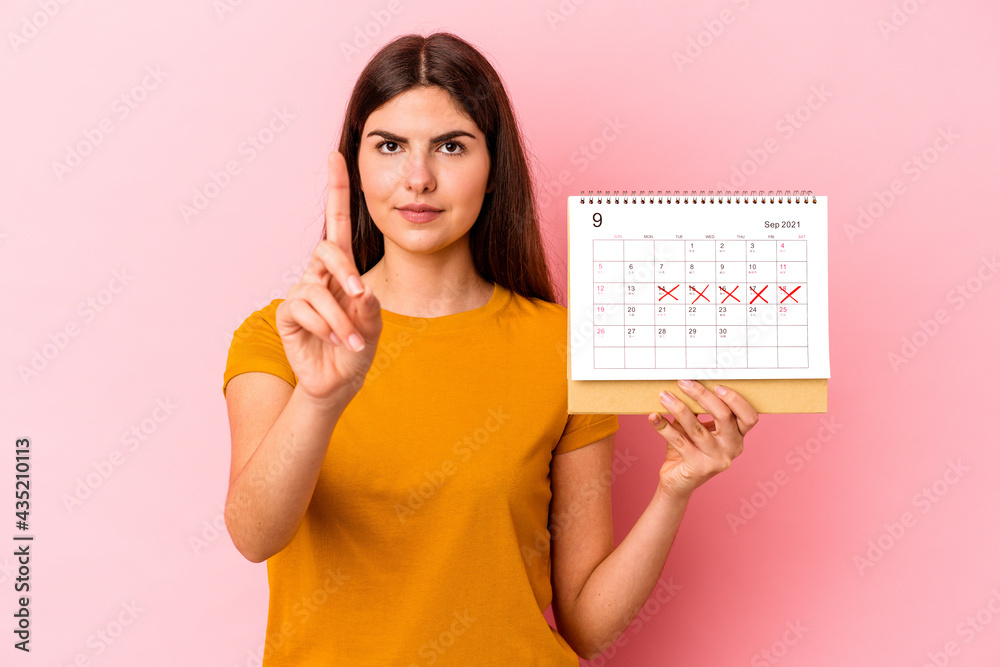 Young caucasian woman holding calendar isolated on pink background showing number one with finger. Young caucasian woman holding calendar isolated on pink background showing number one with finger.