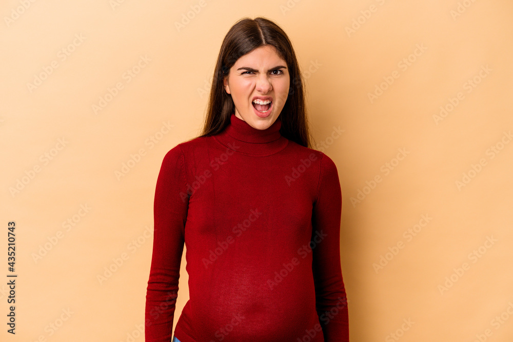 Obraz premium Young caucasian pregnant woman isolated on beige background screaming very angry and aggressive.