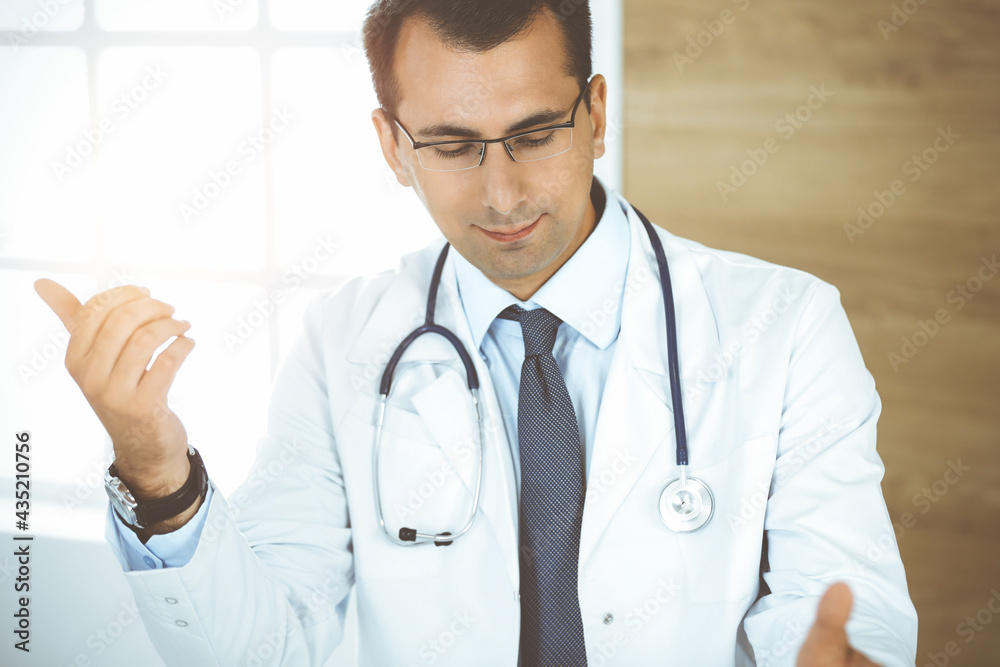 Man-doctor sitting at the desk at his working place and smiling. Perfect medical service in sunny clinic Man-doctor sitting at the desk at his working place and smiling. Perfect medical service in sunny clinic