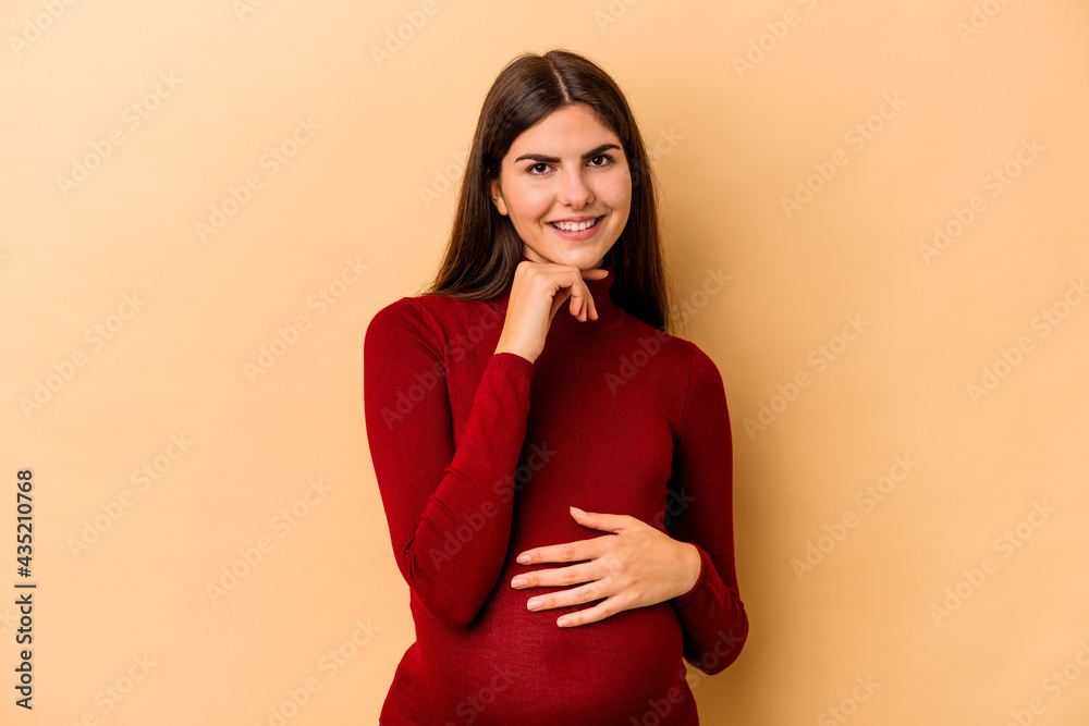 Obraz premium Young caucasian pregnant woman isolated on beige background smiling happy and confident, touching chin with hand.