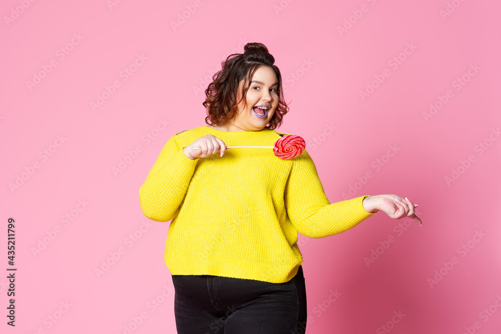 Happy plus size model in casual clothes with big lollipop, fat woman in yellow jumper on pink background Happy plus size model in casual clothes with big lollipop, fat woman in yellow jumper on pink background
