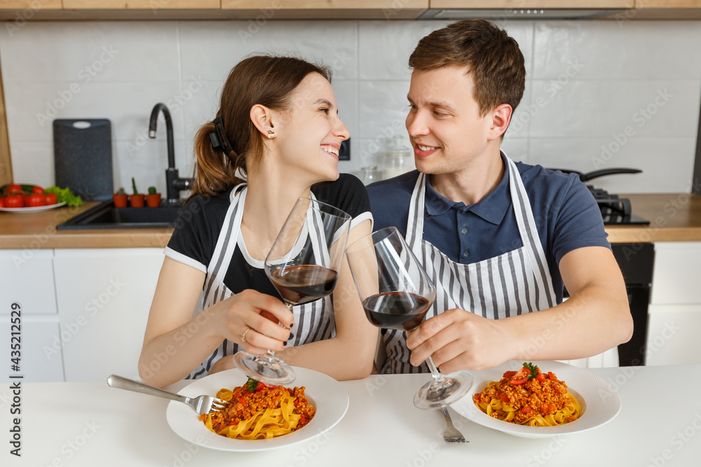 Happy couple in aprons eating bolognese pasta with wine at modern kitchen. Romantic dinner at home. Concept of domestic lifestyle, happy marriage and togetherness